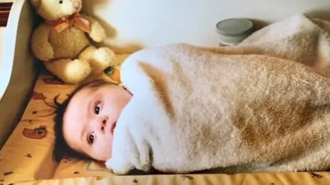 Ellie Goldstein Ellie as a baby wrapped up in a blanket. A teddy bear with a bow around its next is in the corner