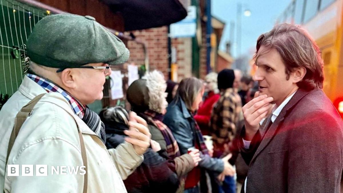 North Norfolk MP Steff Aquarone talking to protesters and residents at the Sheringham Bus Shelter. He is on the right and is wearing a suit and is deep in conversation with a man, standing on the left, who is wearing a coat, a chequered shirt and a flat cap.