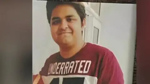 BBC A photograph of Yuvraj Mehta - wearing a maroon-and-white tee shirt - posing for the camera, in a room with white walls