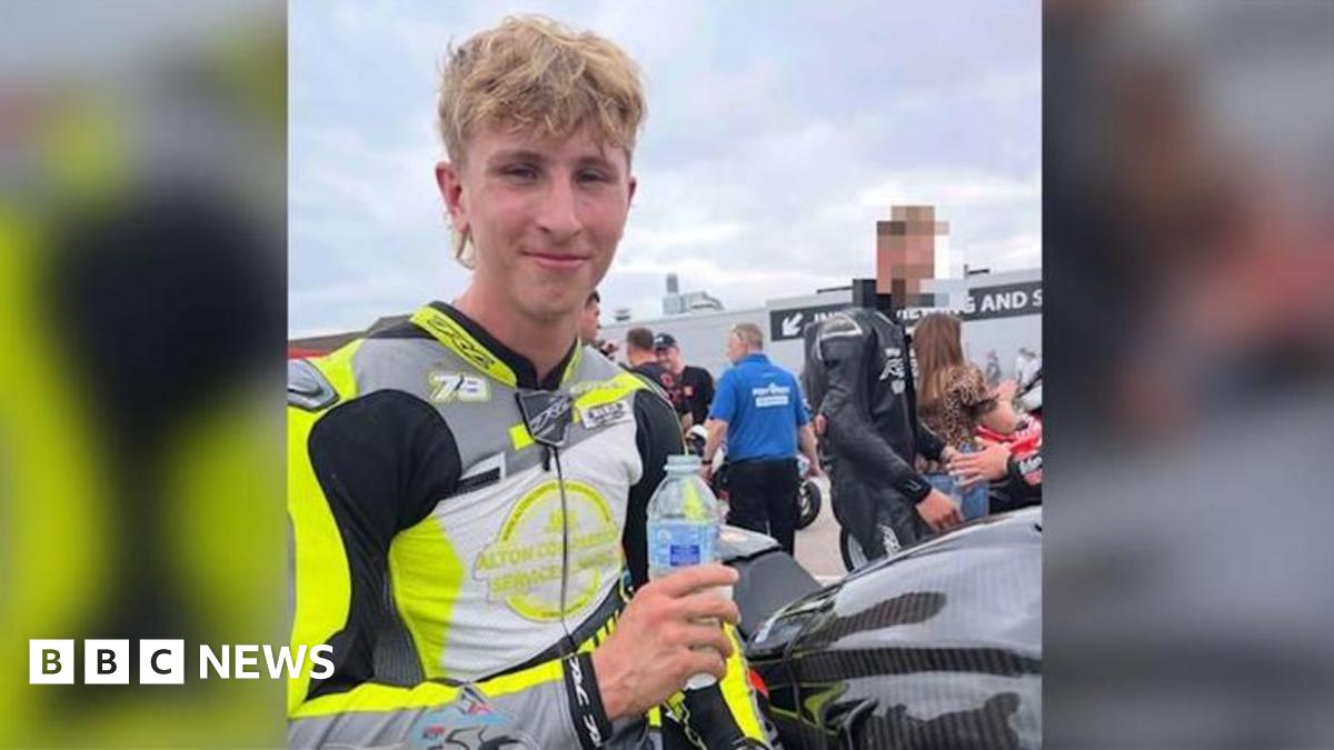 A young blonde man wearing a motorbike jacket and holding a bottle of water.