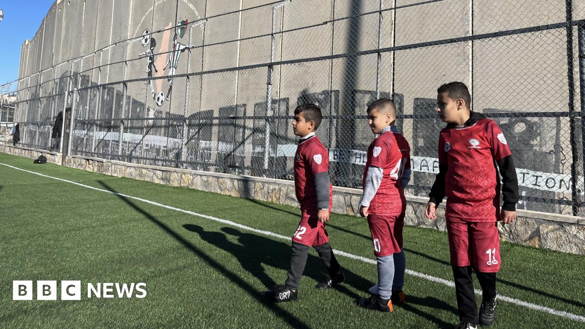 Three Palestinian boys around 10 years old, wearing red football kit, wait in line to take a penalty. They are standing on a green astro turf football pitch with a metal fence behind them. Behind that stands a tall concrete wall