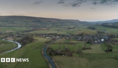 This is an overhead shot of Bainbridge, in the Yorkshire Dales. A river is winding through the left of the shot, the village is on the right, the hills are in the background, a dark and threatening sky above. North Yorkshire Council says the size - and rural nature - of the area it serves adds extra costs to its services.