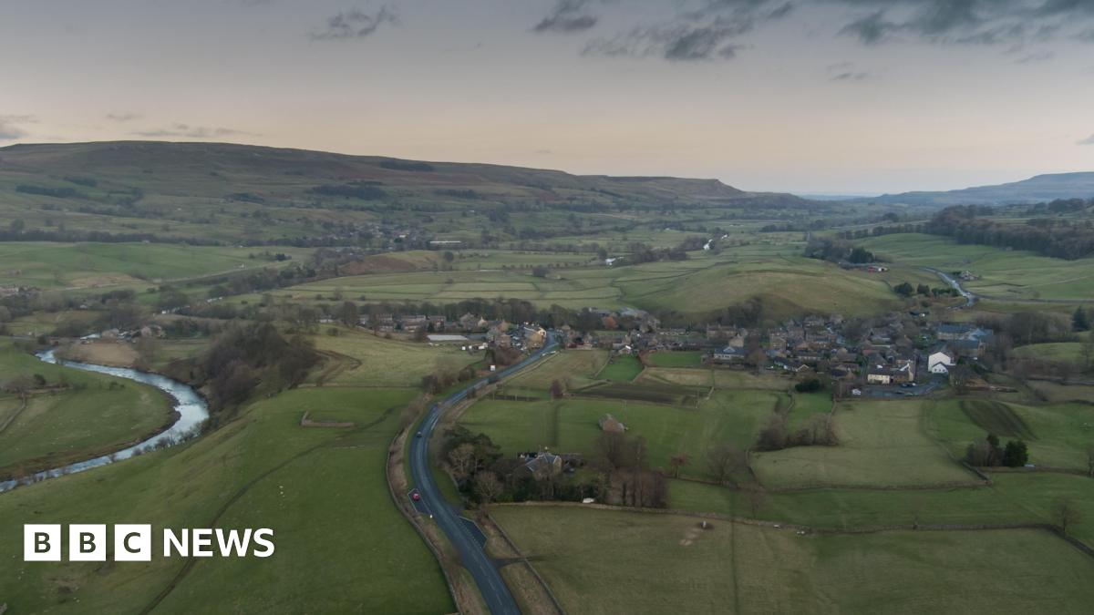 This is an overhead shot of Bainbridge, in the Yorkshire Dales. A river is winding through the left of the shot, the village is on the right, the hills are in the background, a dark and threatening sky above. North Yorkshire Council says the size - and rural nature - of the area it serves adds extra costs to its services.