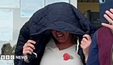 A close-up photo of Rebecca Pinckard walking out of court with her black coat over her head as she tries to cover her face. She appears to be smiling under the coat. She is wearing a cream jumper with a poppy on it.
