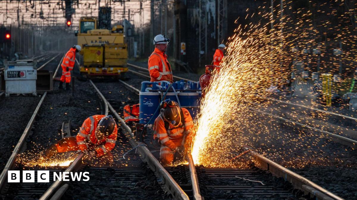 Workers in orange clothing and white hard hats work on a railway line at dusk. They are wearing safety helmets as a jet of bright sparks flies from the track they are using tools on.