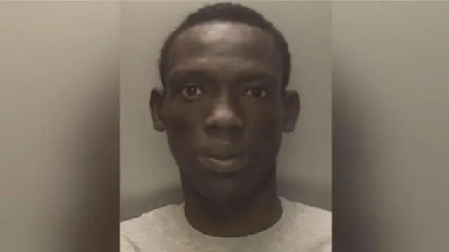 A mugshot of a man with short black hair and wearing a grey jumper, standing against a wall.