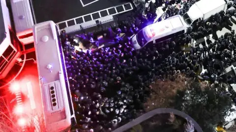 Reuters Thousands of police officers and investigators from South Korea's Corruption Investigation Office gather in front of the entrance to the official residence of impeached South Korean President Yoon Suk Yeol. Several white buses are seen in this shot photographed from above.