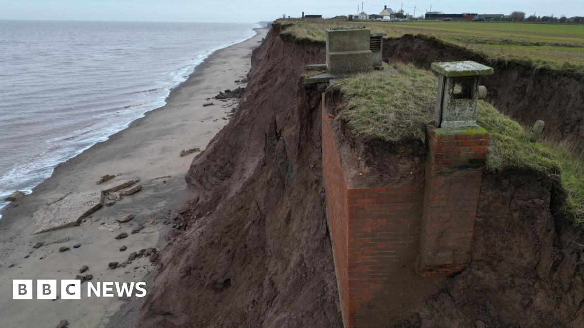 Still shot of the bunker at foot of the cliff. It is leaning into the collapsed cliff face and is surrounded by rock and sand.