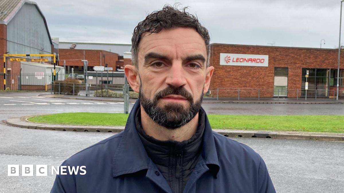 A man with dark hair and a dark beard looking at the camera with a serious expression on his face. He is wearing dark outdoors clothing and standing by the side of the road in front of the main entrance, a red brick building, for Leonardo Yeovil which has a sign reading 'Leonardo' with the company logo