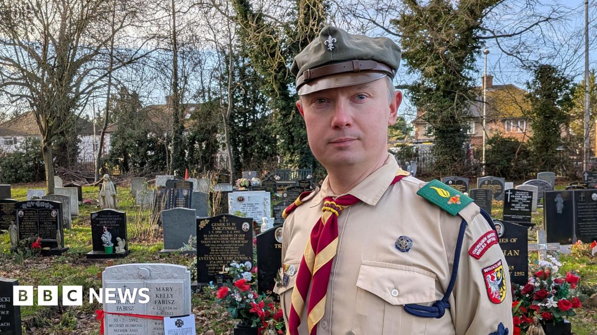 Lukasz Lakomy wearing the Polish Scouting Association uniform and standing in a cemetery. He is wearing a peaked cap in khaki and a light brown shirt with badges. A yellow and red tie is around his neck. Behind him are black, white and grey graves, grass, trees, hedges and beyond that houses.