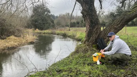 A man in a grey sweatshirt and khaki trousers kneels on a green riverbank next to a tree. He is using equipment from a yellow box to test the water.