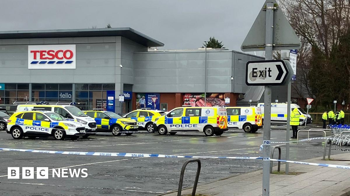 Police vehicles cordon off a Tesco supermarket car park with blue‑and‑white tape as officers stand nearby under a grey sky.