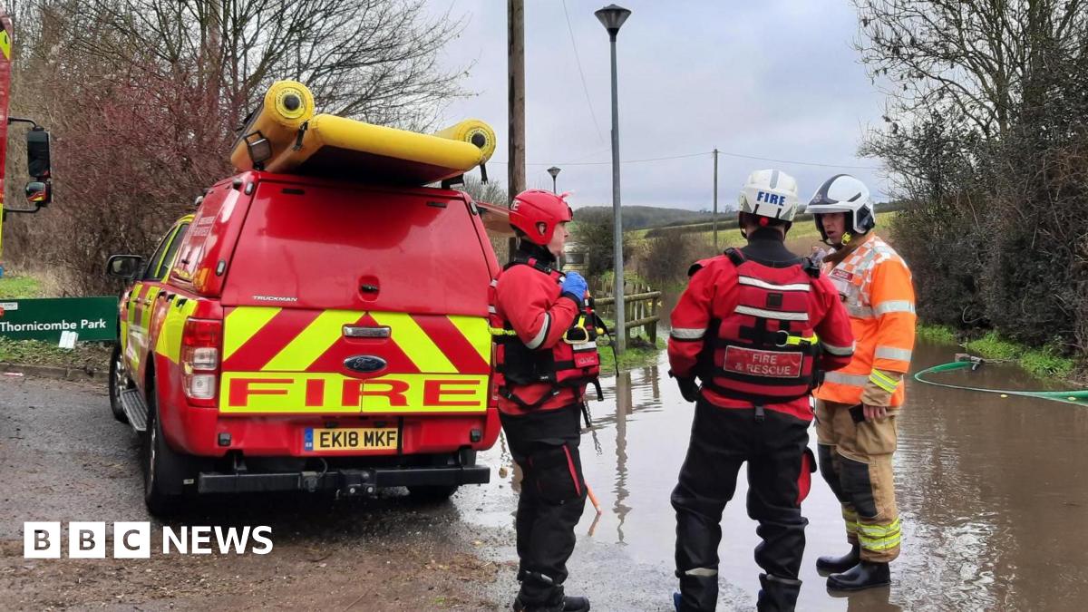 Three firefighters are stood in conversation next to a patch of land flooded with water and a red van with the word 'Fire' printed on the back in large letters. The surroundings are rural, with several bare trees and green fields in the background. To the left of the van is a green sign that reads "Thornicombe Park".