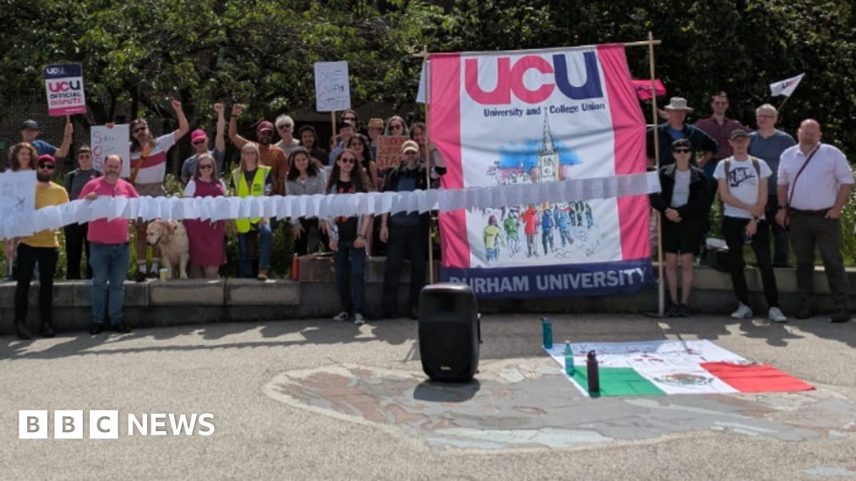 UCU members forming a picket line outside Durham University. Several people in the large group are carrying placards, while two people are supporting a large pink and white UCU banner.