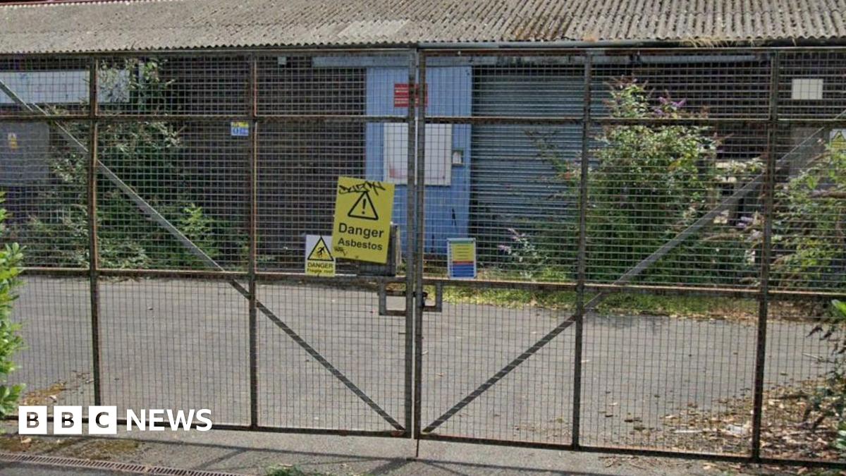 A yellow sign that reads "Danger Asbestos" is pinned to a metal gate. Behind the gate is a metal industrial unit which appears to be dilapidated with vegetation growing in front of it.