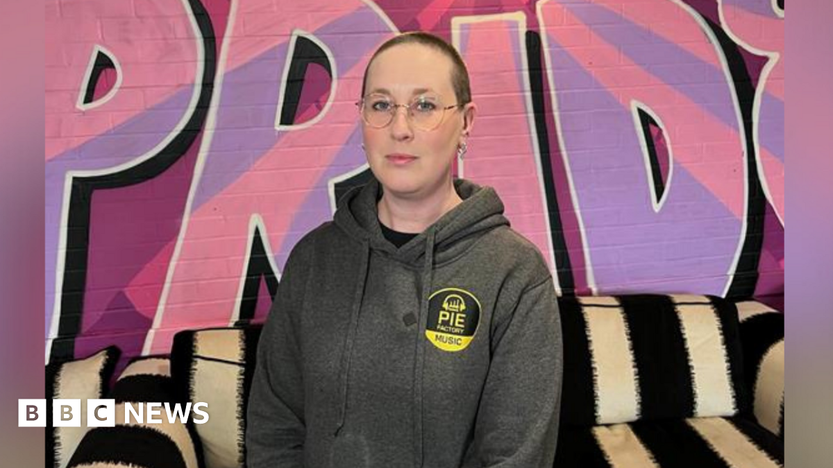 CEO of Pie Factory Music, Zoë Carassik, wearing a grey hoodie and stood against the backdrop of the Pie Music Factory charity headquarters in Ramsgate. 'Pride' is painted in pink and purple on the wall and Zoe is stood in front of a black and white striped sofa.