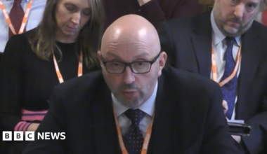 A bald man in black glasses and a black suit with white shirt and black tie. He is sat looking forwards to a person off to the left of the camera. Two people in suits are sat behind him, also facing forwards