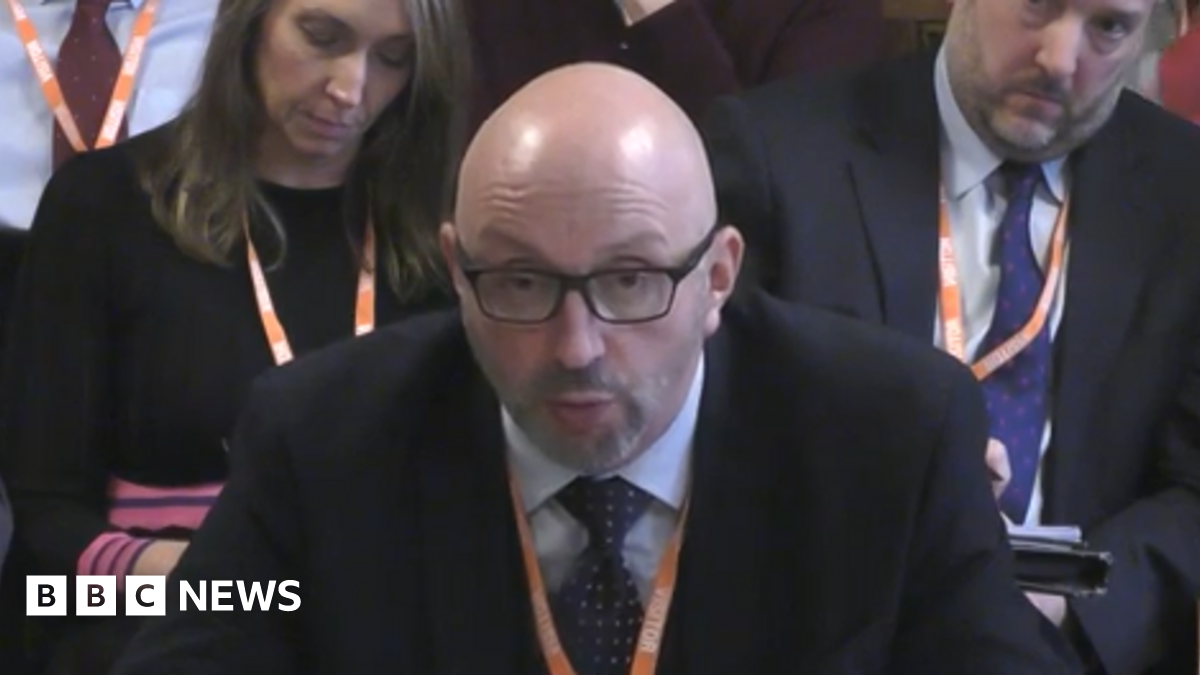 A bald man in black glasses and a black suit with white shirt and black tie. He is sat looking forwards to a person off to the left of the camera. Two people in suits are sat behind him, also facing forwards