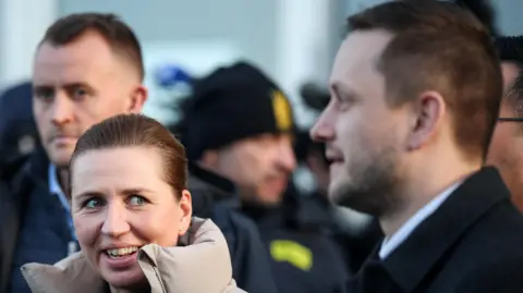 Reuters Danish PM Mette Frederiksen, wearing a puffer jacket with the collar up, smiles and looks towards Greenland PM Jens-Frederik Nielsen, who stands in profile at the right of the picture.