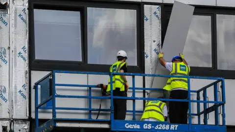 PA Media Three workmen in hi-vis jackets and helmets removing cladding from a building
