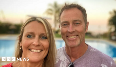 Craig and Lindsay Foreman - they are both standing outside - Lindsay has blond hair and a wide smile - Craig is smiling widely and has brown hair, and a casual light purple t-shirt - a swimming pool and sunset can be seen out of focus behind them