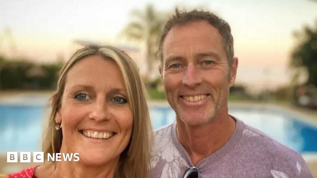 Craig and Lindsay Foreman - they are both standing outside - Lindsay has blond hair and a wide smile - Craig is smiling widely and has brown hair, and a casual light purple t-shirt - a swimming pool and sunset can be seen out of focus behind them