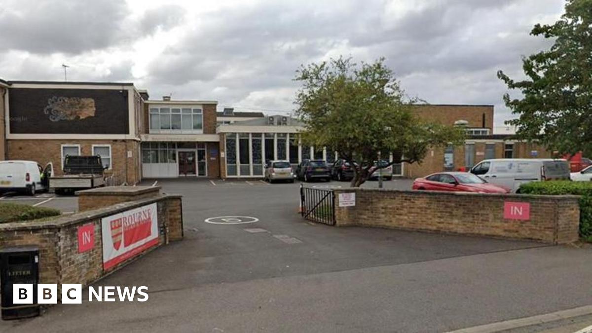 A street view of Bourne Grammar School which shows a brick building with a parking area in front of it. The building has large windows and features a mural of a face on a large dark panel. A wall bordering the building has a banner featuring the word "Bourne" and a red and gold crest. A tree is visible next to a metal gate and the sky is cloudy.