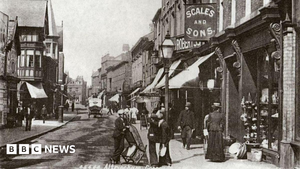 A black-and-white period photograph of George Street in Altrincham from Trafford Council's archive. In the foreground a group of three men in suits can bee seen talking - one of them is holding a rocking chair. A woman in an ankle-length skirt, jacket and hat can be seen looking in a shop window. A horse and cart can be seen further up the street.