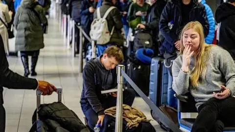 Getty Images Travelers queue with their luggage at Schiphol Airport. One woman is sitting with her phone in her hand, while a man behind her is kneeling next to his bag while reading a book.