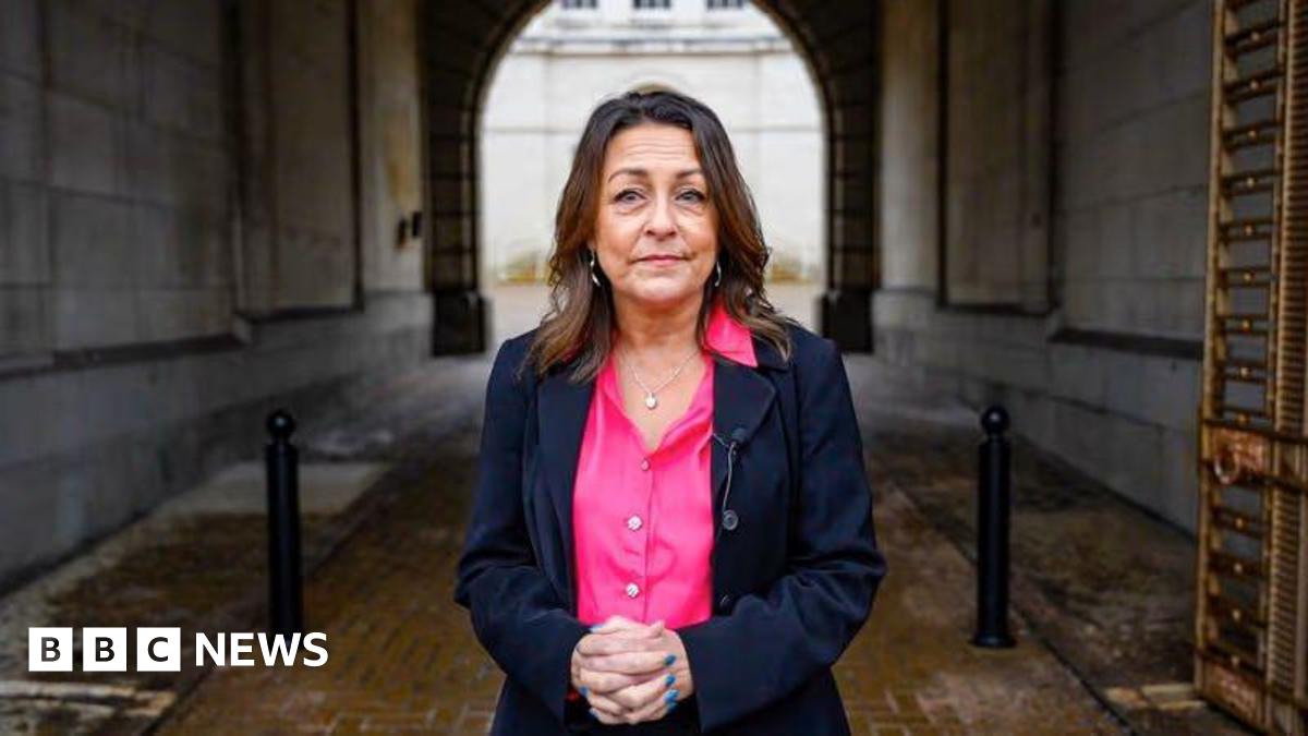 A woman with dark hair wearing a dark suit and pink blouse.
