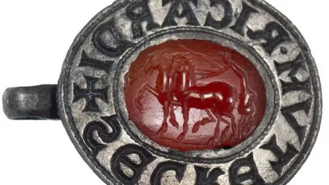 Colchester and Ipswich Museum Service A close-up of an oval silver seal matrix, with a hanging loop on the left side. It is resting on its side, to show the carving of two horses, with upraised legs and a charioteer leaning over them from his chariot with a whip on the oval red gemstone intaglio in the centre. Its thick silver rim is carved with letters. 