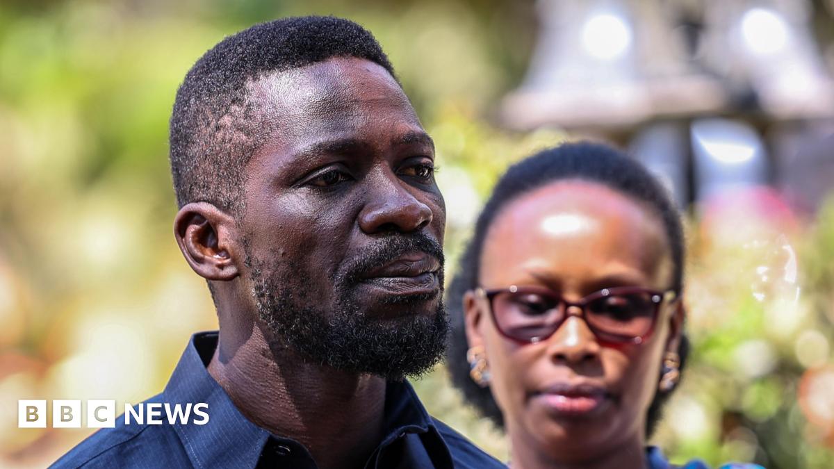 A close-up shot of Bobi Wine looking across the camera. He has a short beard and short hair. His wife is in the background, out of focus.