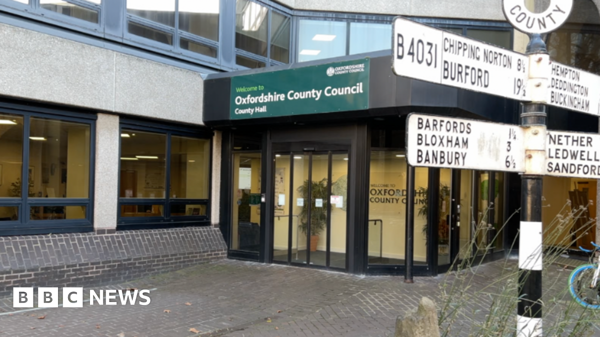Oxfordshire County Council's County Hall in Oxford. The picture shows the front doors of the building, with a green sign above that says "Welcome to Oxfordshire County Council County Hall". There is a black and white sign to the right of the picture pointing out distances to other places in Oxfordshire.