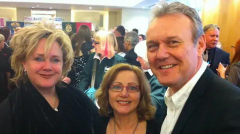 Holly Hedge Sarah Fisher and Anthony Head with Holly hedge founder Pauline Leeson, who is on the middle. 
They are all wearing black and there are people in the background. They are all smiling at the camera.