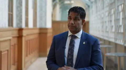 A man wearing a navy blue suit and tie stands in a corridor, looking towards a camera. His hands are clasped together. Glass and wooden panels can be seen in the background behind him.
