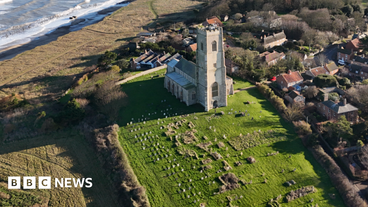 A church viewed from the air, showing its graveyard, fields and the sea beyond, to left, and housing to the right.