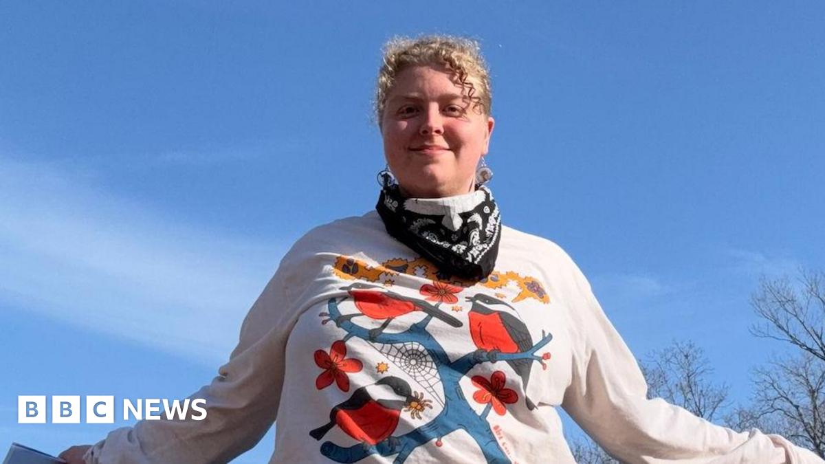 Paradox Mercer looking at camera. She has blonde curly hair. She is wearing a white long-sleeve top with an image of a blue branch with red and dark blue birds on it. There is blue sky in the background with trees. She has a bandanna round her neck.