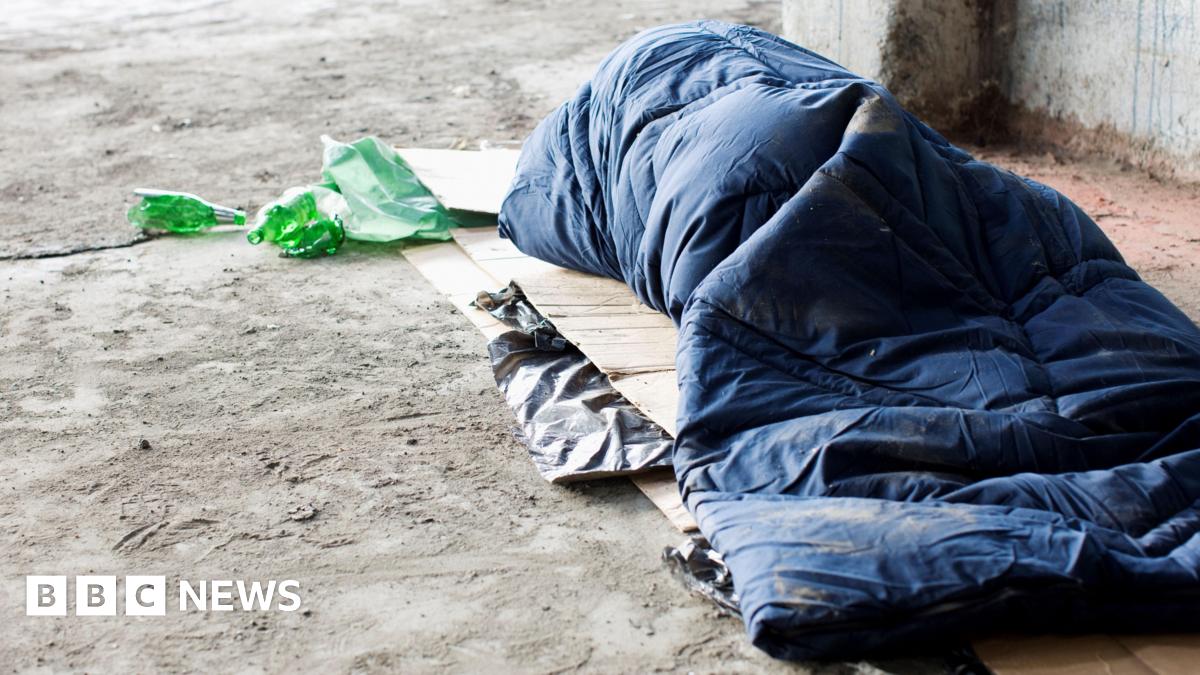 A homeless person sleeps in daylight in a navy blue sleeping bag outdoors on a concrete floor next to a concrete wall. Flattened cardboard boxes are visible underneath the sleeping bag and some green drinks bottles are laid next to the person's head.