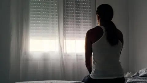 Getty Images A woman wearing a white vest top with her hair tied back sits with her back to the camera, looking out the window.