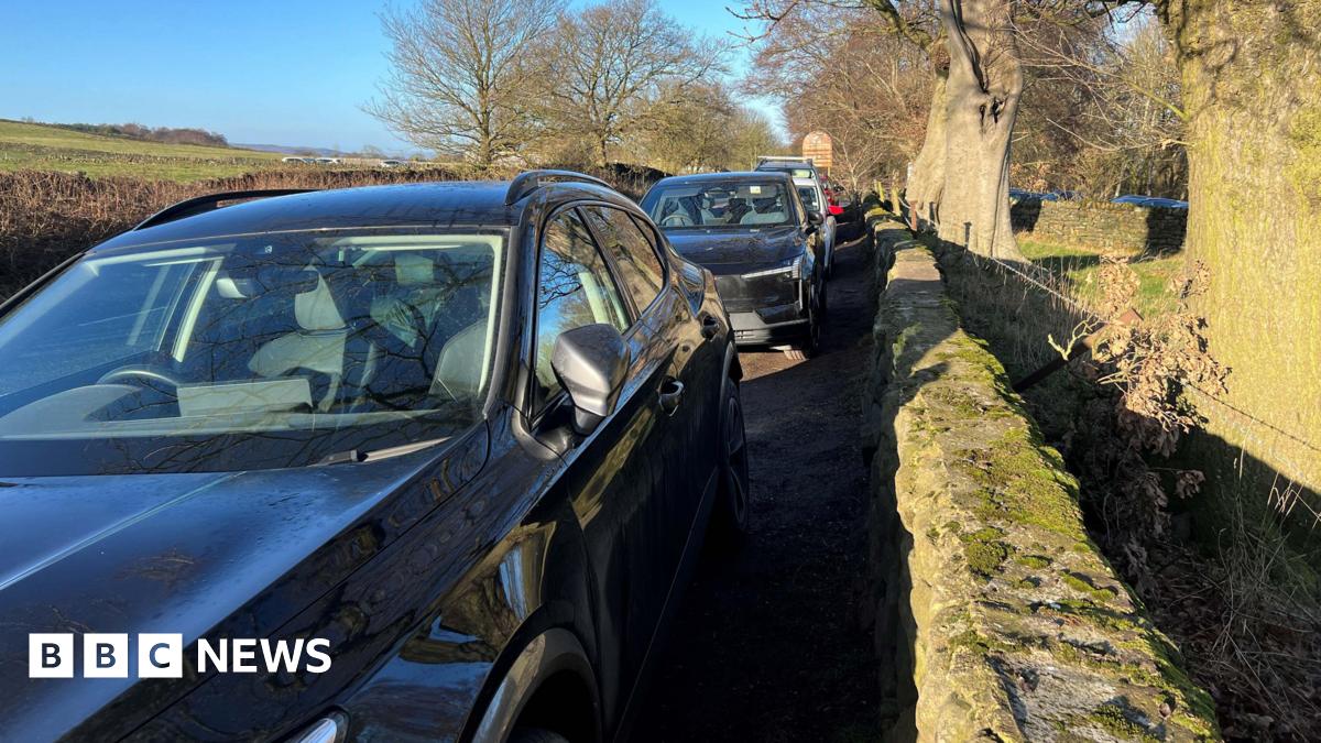 Millie Bobby Brown and David Harbour in character as Eleven and Jim Hopper. Eleven wears red shorts over grey leggings and a grey jumper. Jim Hopper wears a black leather jacket, a black beanie hat and dark trousers.They are speaking to director Frank Darabont on the set of Stranger Things. There is a vintage advertisement posted in the background with debrief on the ground.