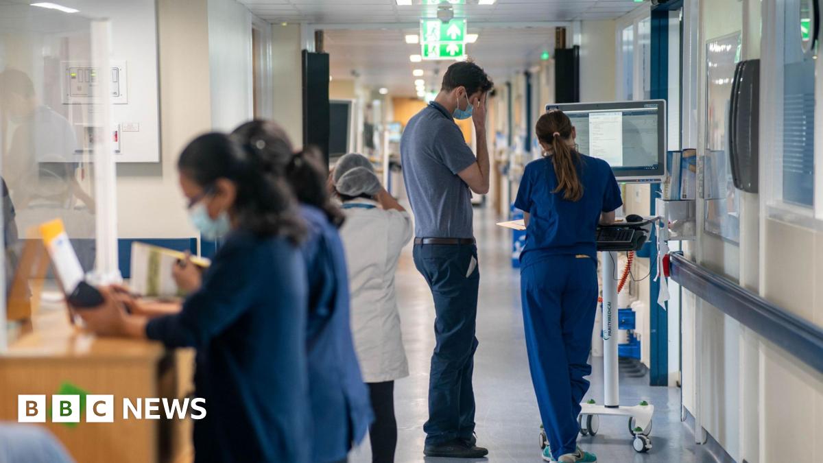 A generic picture of a hospital corridor, two female medical professionals in the foreground leaning on the desk of a nurses' station, an auxiliary walking away beyond them, and what looks like two doctors, a tall man and a shorter woman - in blue scrubs - leaning on a computer on wheels and looking at papers in a folder.
