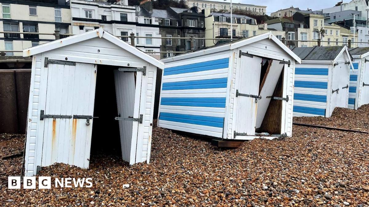 A line of beach huts all scattered across a stony beach and not where they are supposed to be. They are blue and white. The pier can be seen in the back ground.