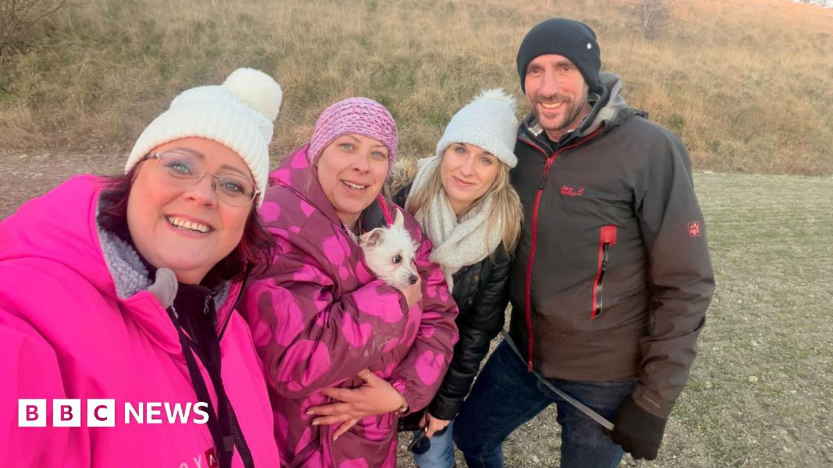 Three women and a man standing in a row on dry grass in warm jackets and woolly hats. Two of the women are in bright pink jackets with a small white dog tucked into one of them. The other woman is in a black leather jacket and white scarf and the man is in a brown waterproof. They are all smiling.