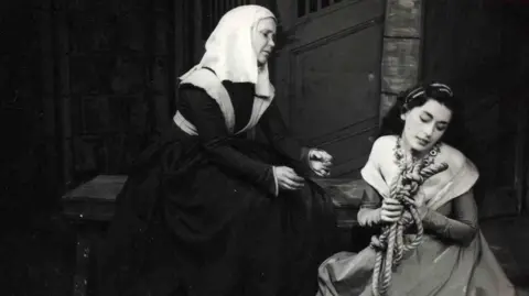 RCS A black and white photo of two women on stage. The woman on the left is sitting on a bench wearing a black dress and a white head covering. The woman on the right is crouched down, wearign a gown and holding a rope.