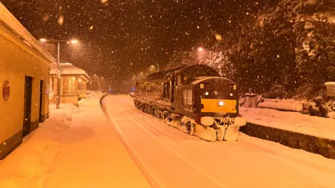 Matt Earnshaw Snow falling heavily during a dark night at a train station, covering both the train station and a locomotive train there