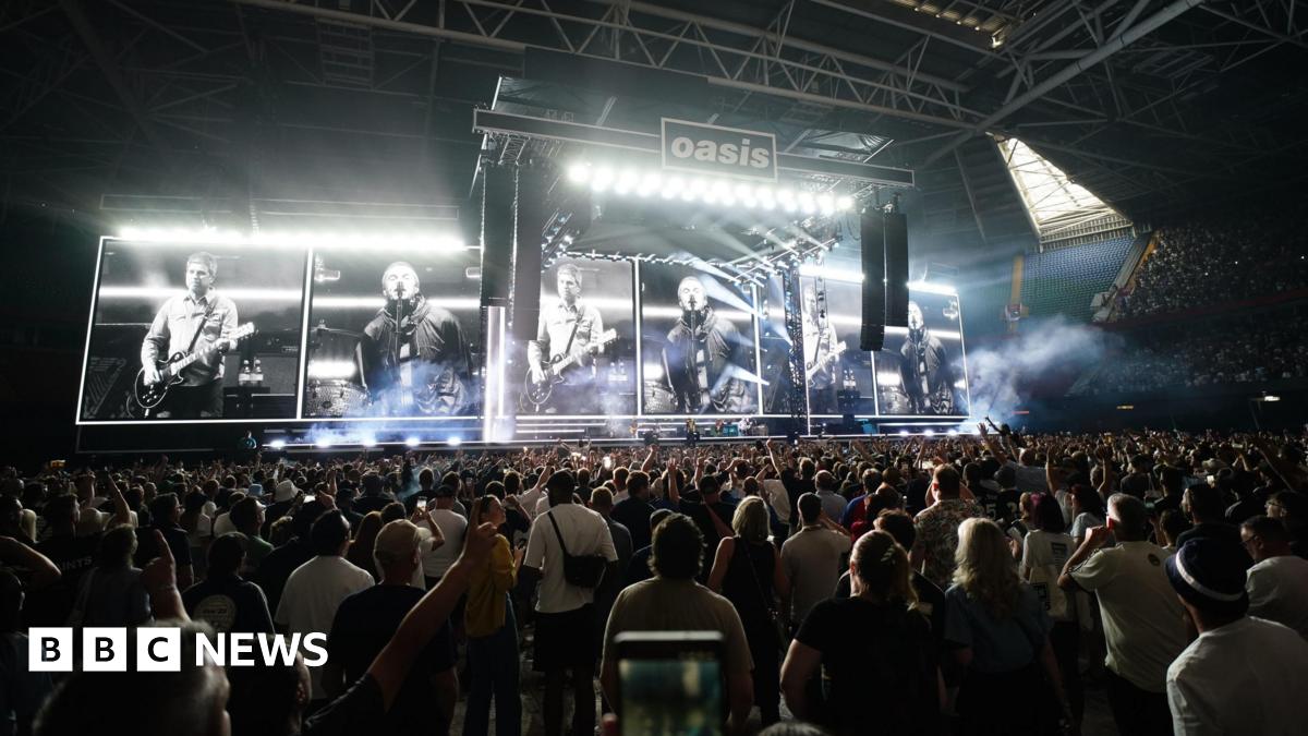 Joe looking at the camera with a straight expression. He has short light brown hair, brown eyes and stubble. He is wearing a navy blue shirt and standing in front of a display case made of large cubes which is blurred in the background.