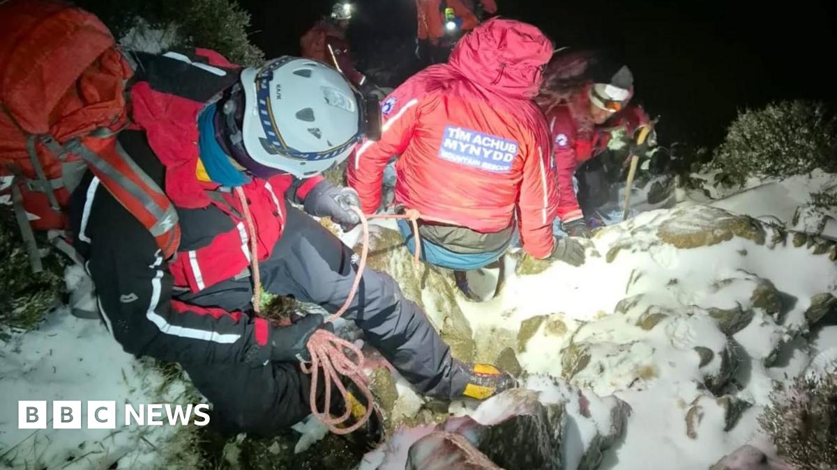 Five volunteers from Ogwen Valley Mountain Rescue descend a snowy mountain in the dark. They wear high visibility red clothing and backpacks and white helmets with torches on.