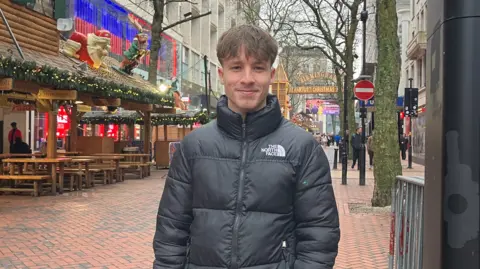A man with brown hair wearing a black puffer jacket smiles at the camera. In the background we can see people walking a long paved street with shops and a covered area with benches. 
