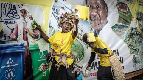 AFP via Getty Images A supporter of Yoweri Museveni in a yellow campaign T-shirt holds flags with Museveni's face on them and is putting up posters with the other.