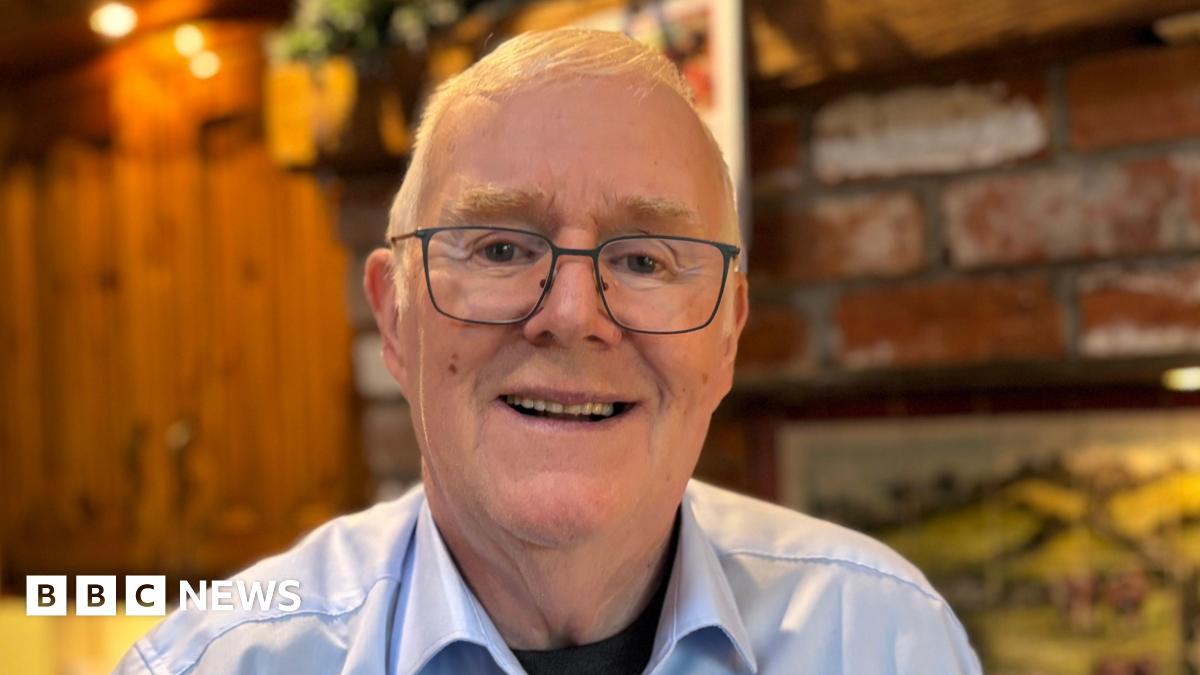 Eugene Reavey. He has short white hair, wearing glasses, a white shirt with black top underneath. Brickwork and cupboards are blurred in the background behind him.